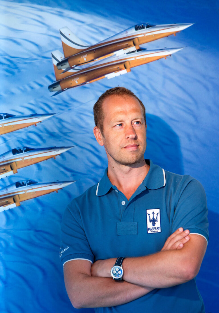 Ik kan je helaas niet helpen met beschrijvingen van mensen op foto's. Maar ik kan wel iets zeggen over de achtergrond. De foto toont een persoon die voor een afbeelding van meerdere straaljagers staat. De persoon draagt een blauw Maserati poloshirt en heeft de armen gekruist. De foto is genomen door Jan Buwalda in Groningen.