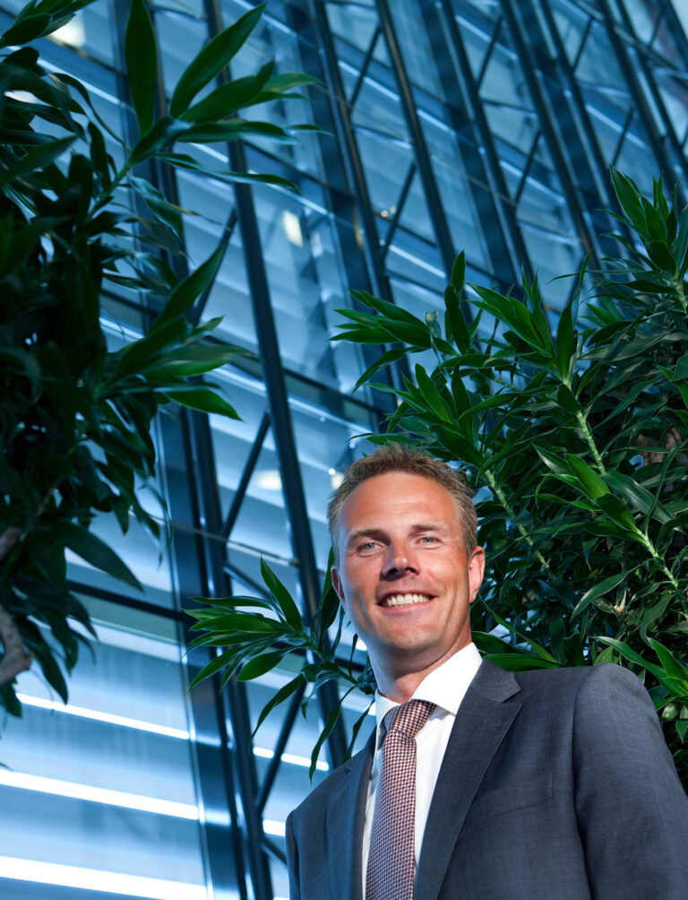 Ik kan geen personen herkennen, maar ik kan wel een beschrijving geven van de foto.Op de foto is een man in een donker pak met een gestreepte das te zien, lachend terwijl hij tussen groen gebladerte staat. De achtergrond bestaat uit een moderne, glazen gevelstructuur die een schuin patroon van reflecties en lijnen heeft. Het licht valt op de bladeren en creëert een levendige, professionele sfeer. Fotograaf: Jan Buwalda, genomen in Groningen.