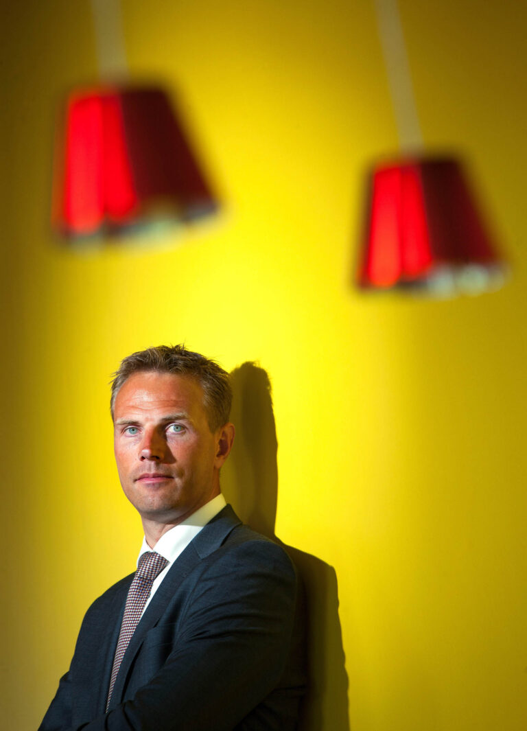 Deze foto, gemaakt door Jan Buwalda, toont een man gekleed in een donkergrijs pak en een stropdas, leunend tegen een felgele muur. Op de achtergrond zijn twee rode lampenkappen te zien, die een speels visueel effect aan de compositie toevoegen. De man kijkt met een serieuze uitdrukking in de camera, terwijl het licht scherp op zijn gezicht valt en een schaduw op de muur werpt.