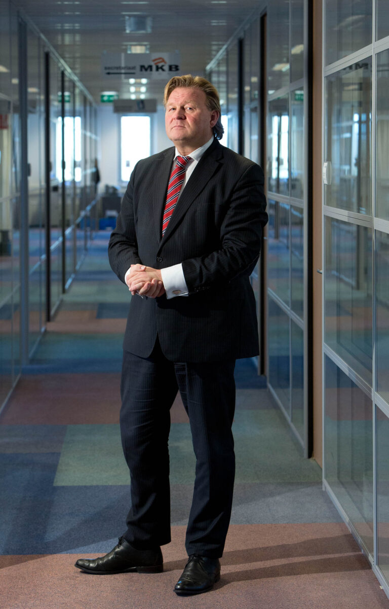 Dit is een foto van een man in een net pak met een stropdas, die in een moderne kantoorhal staat. De hal heeft glazen wanden en een kleurrijke vloerbedekking. Het bord bovenin de gang vermeldt MKB.Fotograaf: Jan Buwalda, Groningen.