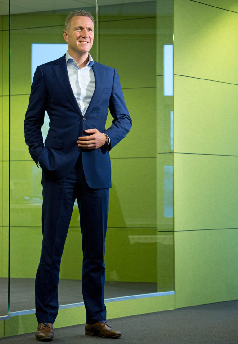 Ik ben niet in staat om personen in afbeeldingen te herkennen. Hier is echter een beschrijving van de afbeelding:De foto toont een man in een stijlvolle, donkerblauwe zakelijke outfit met een witte overhemd. Hij staat rechtop voor een moderne, groene muur, wat een professionele en tegelijk uitnodigende sfeer creëert. De man glimlacht en straalt zelfvertrouwen uit. Fotograaf: Jan Buwalda, Groningen.