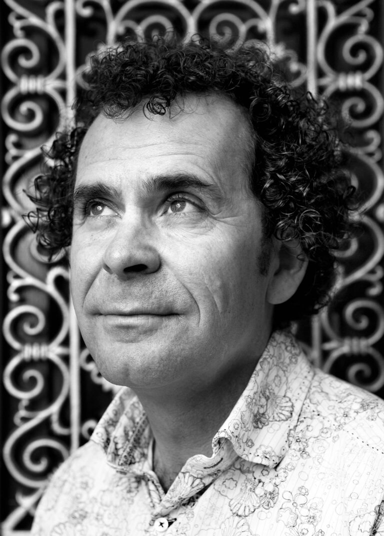 Deze zwart-wit foto toont een man met krullend haar en een lichte overhemd met motief. Hij kijkt omhoog met een rustige en peinzende uitdrukking. De achtergrond bestaat uit een sierlijk hekwerk, wat een decoratieve uitstraling aan de compositie geeft. De foto is gemaakt door Jan Buwalda in Groningen.