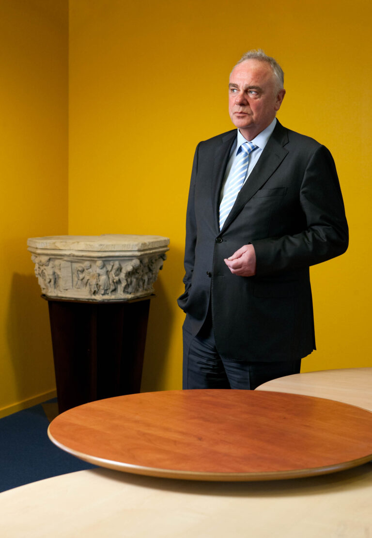 Ik kan de persoon in de afbeelding niet identificeren, maar ik kan wel een beschrijving geven.Op de foto, genomen door Jan Buwalda in Groningen, staat een man in een donkere zakelijke outfit. Hij draagt een blauw gestreepte stropdas. De achtergrond is helder geel en er is een sculptuur te zien. Op de voorgrond bevinden zich ronde houten tafels.Fotograaf: Jan Buwalda.