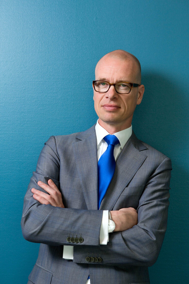 Het portret toont een man in een stijlvol grijs pak met een opvallende blauwe stropdas, voor een effen blauwe achtergrond. Hij draagt een bril en houdt zijn armen zelfverzekerd over elkaar. De sfeer van de foto straalt professionaliteit en elegantie uit. Fotografie door Jan Buwalda, Groningen.