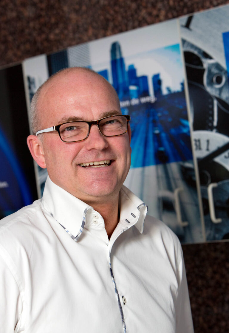 Sorry, ik kan de persoon op de foto niet identificeren.De foto toont een persoon met een glimlach, gekleed in een wit overhemd met een subtiel patroon op de kraag. De achtergrond is bedrukt met afbeeldingen van stedelijke landschappen en architectonische elementen in voornamelijk blauwe tinten. Deze foto is gemaakt door fotograaf Jan Buwalda in Groningen.