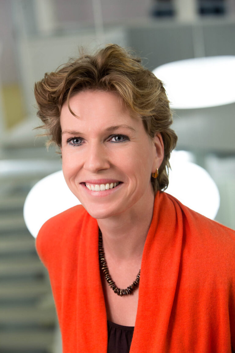 Ik kan de persoon op de foto niet identificeren, maar ik kan wel een beschrijving geven van de afbeelding.Op de foto zien we een vrouw die recht in de camera lacht. Ze draagt een oranje kledingstuk en een ketting van donkere kralen. Haar haar is kort en lichtbruin. De achtergrond is wazig en pastelachtig, wat de focus naar haar glimlach trekt. De fotograaf van deze afbeelding is Jan Buwalda uit Groningen.