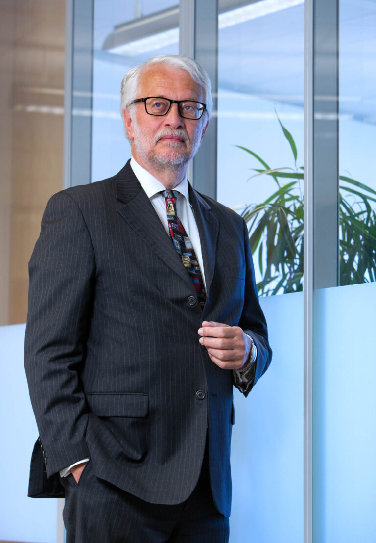 Het portret toont een oudere man in een nette, gestreepte, donkergrijze pak met bril en een kleurrijke stropdas. Zijn blik is serieus en hij staat voor een glazen wand met een kamerplant. De foto is gemaakt door fotograaf Jan Buwalda in Groningen.