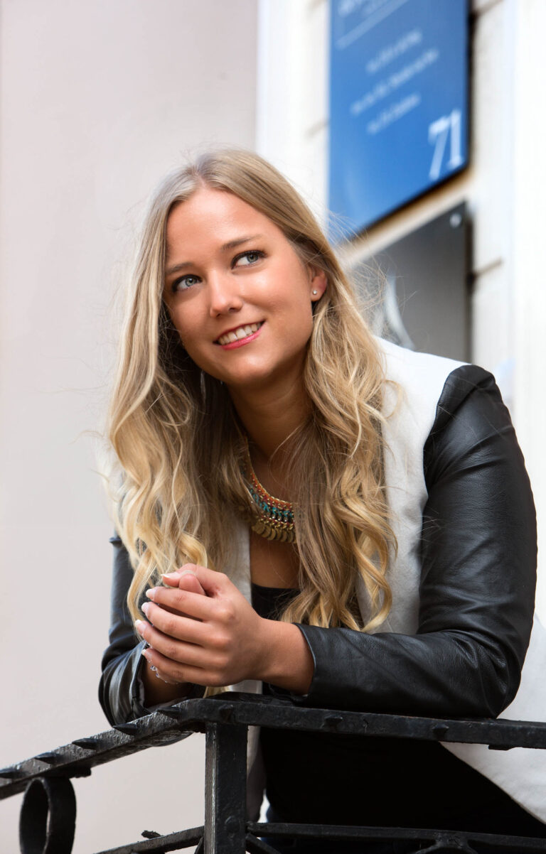 Deze foto, genomen door Jan Buwalda in Groningen, toont een jonge vrouw met lang blond haar en een vriendelijke glimlach. Ze leunt op een metaal hek, gekleed in een zwart-witte jas en een kleurrijke ketting. Op de achtergrond is een gevel te zien met een blauwe plaat. De sfeer van de foto is ontspannen en spontaan.