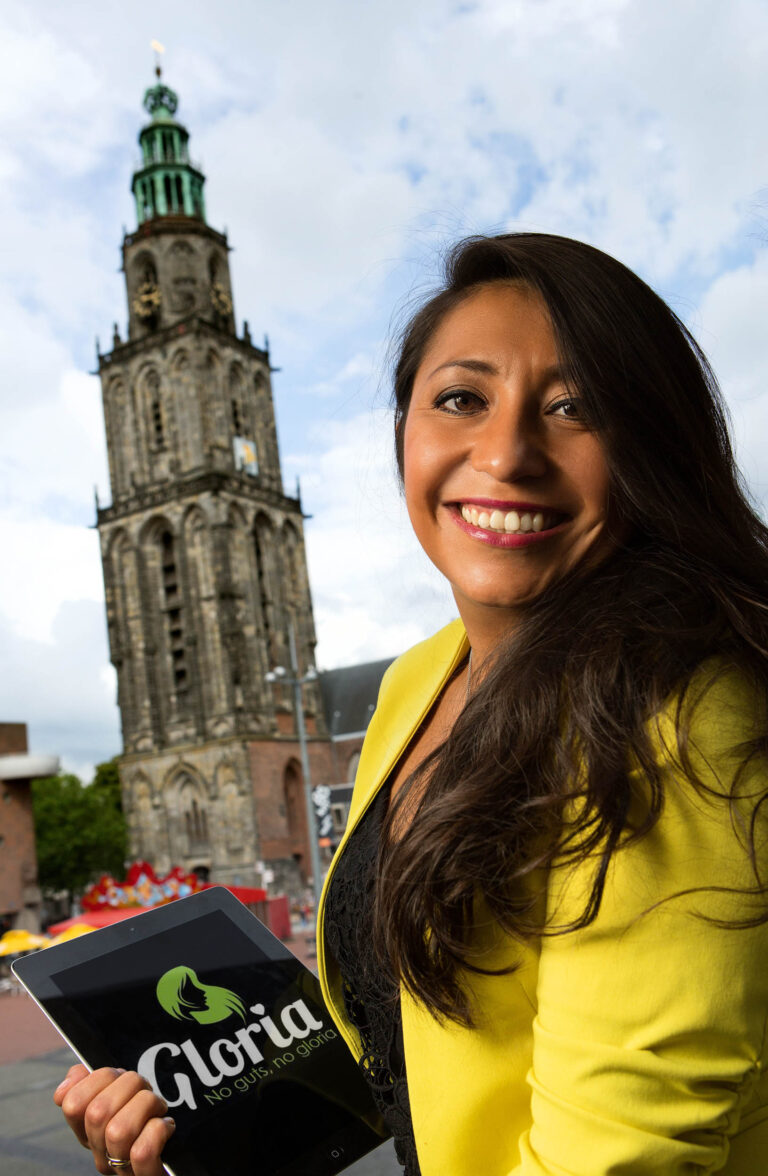 Het beeld toont een glimlachende vrouw die een tablet vasthoudt met het logo van Gloria erop. Ze draagt een felgele blazer en staat voor een herkenbare locatie in Groningen: de Martinitoren. De toren is het prominente middelpunt op de achtergrond, tegen een bewolkte hemel. De foto straalt een moderne en levendige sfeer uit. Fotografie door Jan Buwalda, Groningen.