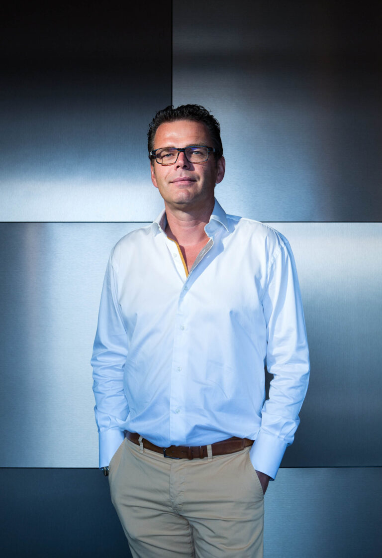 Ik kan de persoon op de foto niet identificeren, maar ik kan wel een beschrijving geven van de afbeelding.De foto toont een man die gekleed is in een witte blouse en beige broek. Hij draagt een bril en heeft donker haar. De achtergrond is een metallic, industrieel paneel dat zorgt voor een moderne uitstraling. De belichting laat een subtiel spel van licht en schaduw zien, wat diepte geeft aan de foto. Fotograaf: Jan Buwalda, Groningen.