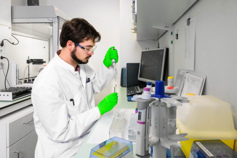 I'm sorry, ik kan de mensen op de foto niet identificeren. Maar ik kan wel een beschrijving geven van de afbeelding.Op de foto zien we een laboratoriumomgeving waar een medewerker in witte labjas en veiligheidsbril bezig is met een pipet. De medewerker draagt groene handschoenen en werkt geconcentreerd aan een werkblad met verschillende labinstrumenten en reageerbuizen. Op de achtergrond staat een computer met verschillende papieren en instrumenten. De foto is genomen door Jan Buwalda in Groningen.