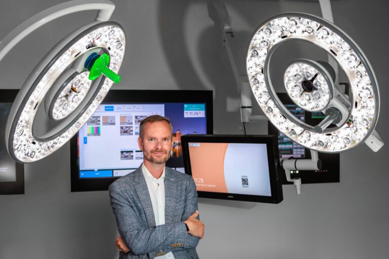 In deze foto zien we een persoon in een moderne medische omgeving, gefotografeerd door Jan Buwalda in Groningen. De persoon heeft zijn armen over elkaar en staat voor enkele high-tech chirurgische lampen en schermen die informatie en beelden weergeven. De ruimte is goed verlicht, en de technologie op de achtergrond suggereert dat het een geavanceerde medische faciliteit is. De algehele sfeer is professioneel en innovatief.