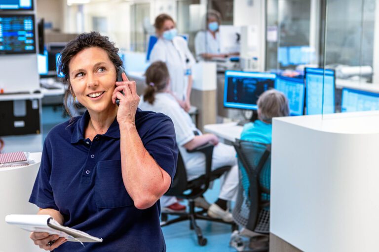 Een vrouw staat in een moderne medische omgeving met een telefoon aan haar oor, glimlachend terwijl ze notities vasthoudt. Op de achtergrond zijn medische professionals te zien die bezig zijn met hun werk aan computers. De ruimte heeft een georganiseerde en efficiënte uitstraling, wat de professionele atmosfeer benadrukt. Foto door Jan Buwalda, genomen in Groningen.