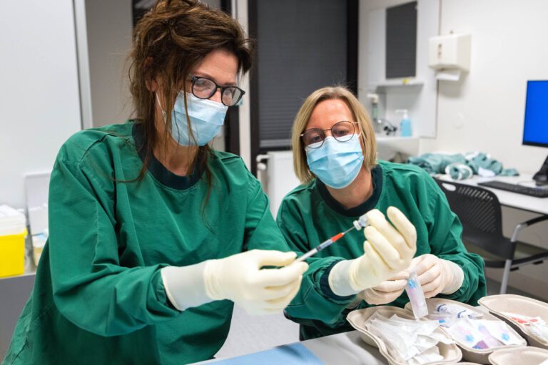 In deze foto, genomen door Jan Buwalda in Groningen, zijn twee medisch professionals te zien die beschermende kleding, handschoenen en mondkapjes dragen terwijl ze werkzaam zijn in een klinische omgeving. Ze zijn betrokken bij het klaarmaken van injecties, omringd door diverse medische benodigdheden op een tafel in een heldere, goed uitgeruste kamer. Hun geconcentreerde houdingen suggereren dat ze zorgvuldig en gedetailleerd werk verrichten.