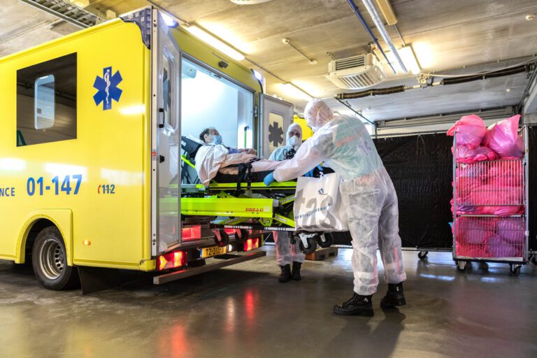 Dit beeld toont medisch personeel in beschermende kleding dat bezig is om een patiënt in een ambulance te plaatsen. De ambulance, geel van kleur, staat geparkeerd in een moderne, goed verlichte garage of oprit. Het personeel is volledig uitgerust met gezichtsmaskers en beschermende pakken, en de patiënt ligt op een brancard met een masker op. Aan de zijkant van de ambulance staat het nummer 01-147 en er zijn rolcontainers met roze zakken in de achtergrond. Foto door Jan Buwalda, genomen in Groningen.