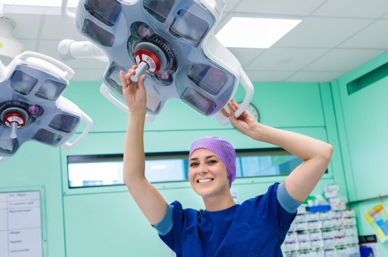 Dit is een foto van een vrolijke zorgmedewerker in een operatiekamer. Ze draagt een paarse operatiemuts en een blauwe operatiejas terwijl ze een chirurgische lamp positioneert. De kamer heeft een mintgroene kleur met kasten op de achtergrond. De sfeer is professioneel en efficiënt. Foto door Jan Buwalda, Groningen.