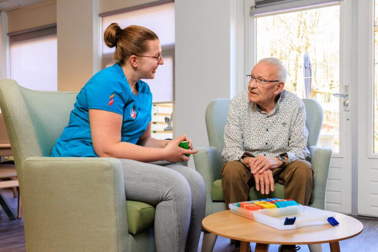 Ik kan helaas geen namen van mensen in foto's verstrekken. Hier is een beschrijving zonder de namen:In een lichte en uitnodigende kamer zit een verpleegkundige in een blauwe uniform lachend tegenover een oudere man. Ze lijken in een vriendelijk gesprek verwikkeld te zijn. Op het tafeltje tussen hen staan kleurrijke doosjes, mogelijk medicatiedoosjes. De inval van het zonlicht door de grote ramen zorgt voor een warme en uitnodigende sfeer in de ruimte. Foto door Jan Buwalda, Groningen.