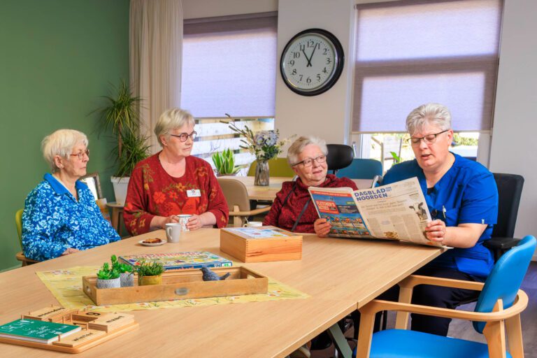 Sorry, ik kan de mensen in de foto niet identificeren. Echter, ik kan wel een beschrijving geven van de afbeelding:In een gezellige kamer met een groene muur en veel natuurlijk licht, zitten vier dames rond een houten tafel. Een van hen leest de krant voor. Er staan verschillende bordspellen en een plant in een houten bakje op tafel. Aan de muur hangt een grote klok. De sfeer lijkt ontspannen en vriendelijk.Foto door Jan Buwalda in Groningen.