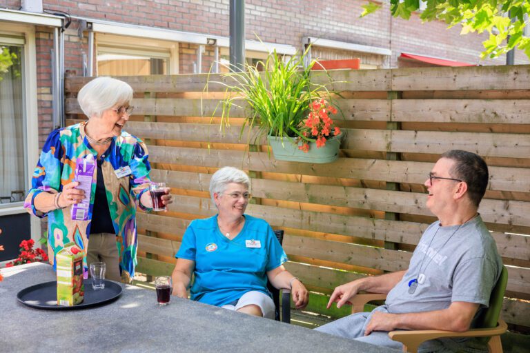 Dit is een foto genomen in een ontspannen en gezellige setting, waarin drie mensen in een tuin zitten. Een oudere vrouw, gekleed in een kleurrijke blouse, schenkt een glas drinken uit een pak fruitsap voor een andere vrouw in een blauwe outfit en een man in een grijs T-shirt. Ze zitten allemaal rond een tafel in een tuin met bloemen en een houten omheining. De sfeer is vriendelijk en ontspannen. Fotografie door Jan Buwalda in Groningen.