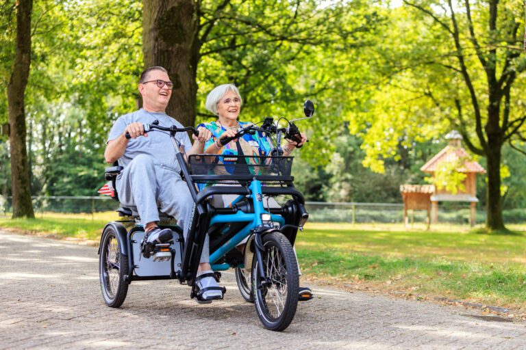 Een oudere vrouw en man genieten samen van een rit op een driewielige fiets in een groene parkachtige omgeving. De zon schijnt door de bladeren van de bomen, wat zorgt voor een warme, vriendelijke sfeer. Ze lijken ontspannen en vrolijk terwijl ze over het verharde pad fietsen. Deze foto is gemaakt door Jan Buwalda in Groningen.