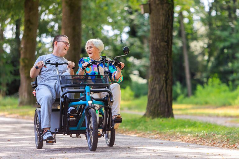 In deze foto, genomen door Jan Buwalda in Groningen, zien we een vrolijk stel ontspannen genietend van een fietstocht op een duofiets. Ze rijden langs een groene, bosrijke omgeving en lachen terwijl ze het pad volgen. De zon werpt zachte schaduwen op de grond, wat bijdraagt aan de serene en blije sfeer van het moment.