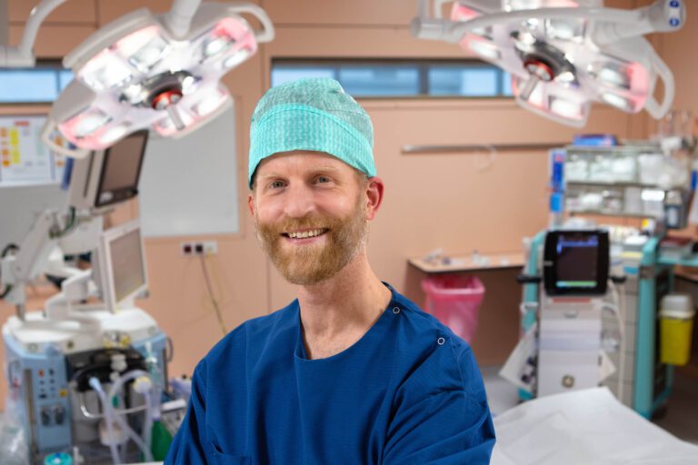 Dit is een foto van een medisch professional in een operatiekamer. De persoon draagt blauwe medische kleding en een groene operatiemuts. Op de achtergrond zijn medische apparatuur en verlichtingsarmaturen zichtbaar, die typerend zijn voor een operatiekamer. De sfeer is klinisch en georganiseerd. Fotograaf: Jan Buwalda, Groningen.