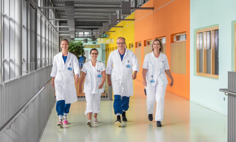 In deze foto genomen door Jan Buwalda in Groningen, zien we vier medisch personeel in witte uniformen die samen door de gang van een moderne zorginstelling lopen. De gang heeft kleurrijke muren in tinten zoals oranje en mintgroen, wat een levendige sfeer creëert. Het licht stroomt overvloedig binnen door de grote ramen aan de linkerkant, en er is een ontspannen en vriendelijke sfeer onder het personeel, wat bijdraagt aan een positieve werkplek.