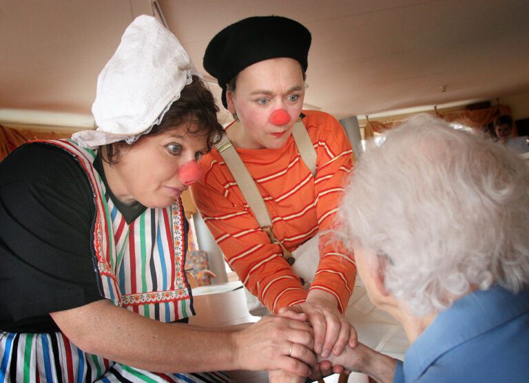 Deze foto toont twee personen verkleed als clowns in kleurrijke outfits en met rode neuzen, die vriendelijk in gesprek zijn met een oudere persoon. Eén clown draagt een wit gehaakt mutsje en een gestreepte schort, terwijl de ander een zwarte baret en een oranje gestreepte trui draagt. Ze lijken zorgzaam en betrokken terwijl ze de oudere persoon vasthouden en met aandacht kijken. De sfeer is warm en vriendelijk. Foto door Jan Buwalda.