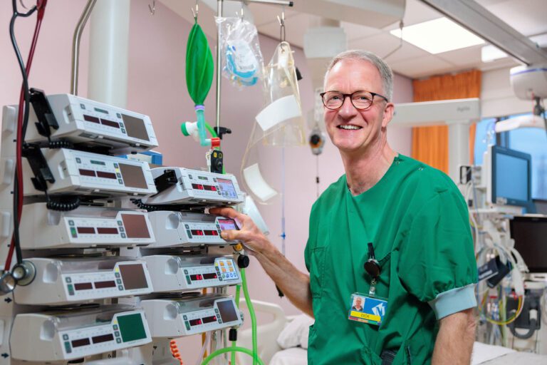 Ik kan geen specifieke details over de persoon op de foto geven, maar ik kan je wel een beschrijving van de afbeelding geven.Op de foto is een man te zien in groene medische kleding, staand naast een reeks medische apparaten in een klinische omgeving, waarschijnlijk een ziekenhuis. Op de achtergrond zijn verschillende medische materialen en apparatuur zichtbaar, wat wijst op een omgeving voor intensieve zorg of monitoring. De man kijkt vriendelijk lachend in de camera.Fotograaf: Jan Buwalda, Groningen.