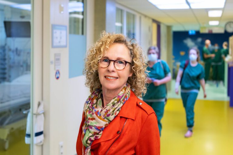 De foto toont een vrouw met blond krullend haar en een rode jas, vriendelijk glimlachend in de camera. Ze draagt een kleurrijke sjaal en een bril. Op de achtergrond is een ziekenhuisomgeving zichtbaar met meerdere medisch personeel in groene uniformen en mondmaskers. De opname is helder en levendig, wat een professionele en zorgzame sfeer uitstraalt. Fotograaf: Jan Buwalda, Groningen.
