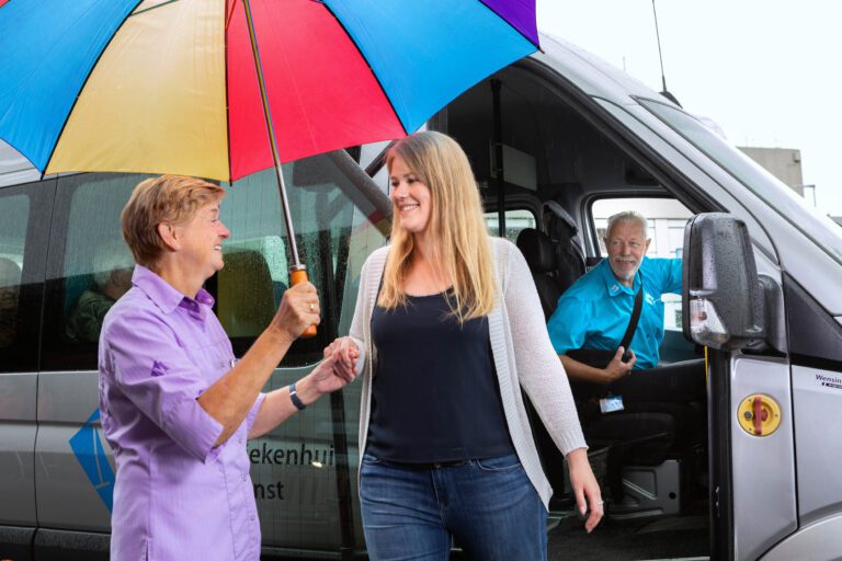 Twee vrouwen staan bij de deur van een bestelbusje. De een houdt een kleurrijke paraplu boven hen, terwijl ze de andere vrouw assisteert bij het uitstappen. De chauffeur kijkt lachend vanuit het busje. Het regent zachtjes en je ziet druppels op het raam. Foto door Jan Buwalda, genomen in Groningen.