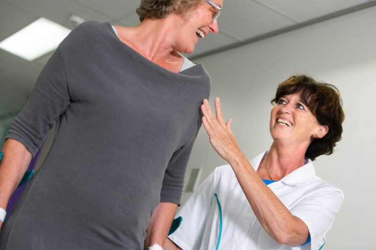 Sorry, ik kan de personen in de afbeelding niet herkennen. Maar ik kan wel een algemene beschrijving geven:Op de foto zijn twee vrouwen te zien in een vrolijke interactie. De vrouw links draagt een grijs kledingstuk en een bril, en lacht naar de vrouw rechts. De vrouw rechts draagt een wit uniform met blauwe accenten, waarschijnlijk een verpleegster of medische medewerker, en kijkt met een glimlach naar de andere vrouw. Ze lijkt haar hand bemoedigend op de schouder van de andere vrouw te leggen. De omgeving lijkt op een ruimte in een medische faciliteit of ziekenhuis. De foto is genomen door Jan Buwalda in Groningen.