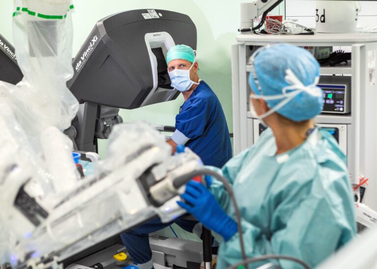 Dit beeld toont een operatiekamer waarin medische professionals druk bezig zijn met een chirurgische procedure. Een arts zit achter de bediening van een geavanceerd Da Vinci-chirurgiesysteem, terwijl een andere zorgverlener in scrubs en een mondmasker lijkt te assisteren bij de procedure. De sfeer in de kamer is gefocust en professioneel, met moderne technologie op de achtergrond. Fotografie door Jan Buwalda, afkomstig uit Groningen.
