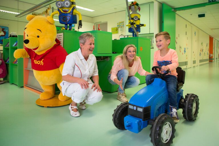Ik zie een vrolijke scène in een kinderziekenhuis. Een jongen lacht terwijl hij op een blauwe speelgoedtractor rijdt. Naast hem zitten een verpleegster en een vrouw op hun hurken en glimlachen naar de jongen. Op de achtergrond staat een groot Winnie de Poeh-beeld en zijn er felgekleurde kasten met speelgoed te zien. De sfeer is vriendelijk en ondersteunend, met een heldere en kleurrijke omgeving die de kinderen op hun gemak probeert te stellen.Fotograaf: Jan Buwalda, Groningen.