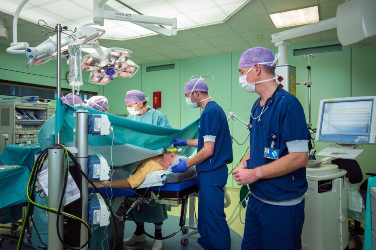 Dit beeld toont een operatiescène in een ziekenhuis, met meerdere medisch specialisten die bezig zijn in een operatiekamer. De sfeer is geconcentreerd en professioneel, met helder chirurgisch licht dat de ruimte verlicht. De chirurgen en het medisch team dragen blauwe en groene scrubs, gezichtsmaskers en paarse haarnetjes. Diverse medische apparatuur en monitors zijn zichtbaar, wat een beeld geeft van de moderne technologische setting waarin de professionals werken. Deze foto is gemaakt door Jan Buwalda in Groningen.