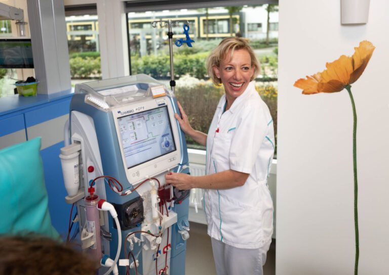 I'm sorry, but ik kan niet zeggen wie er op deze foto staat. Wel kan ik de scène beschrijven.Op de foto zien we een zorgverlener in een witte verpleegkundigenuniform, werkend met een medisch apparaat in een lichte en moderne zorgomgeving. De zorgverlener kijkt ontspannen en tevreden naar iets of iemand buiten de opname. Links van hen staat een dialyse-apparaat met diverse slangen en een scherm met daarop medische informatie. Op de achtergrond is er daglicht dat door het raam naar binnen valt, en naast de zorgverlener is een muurdecoratie met een afbeelding van een bloem te zien.Fotograaf: Jan Buwalda, Groningen.