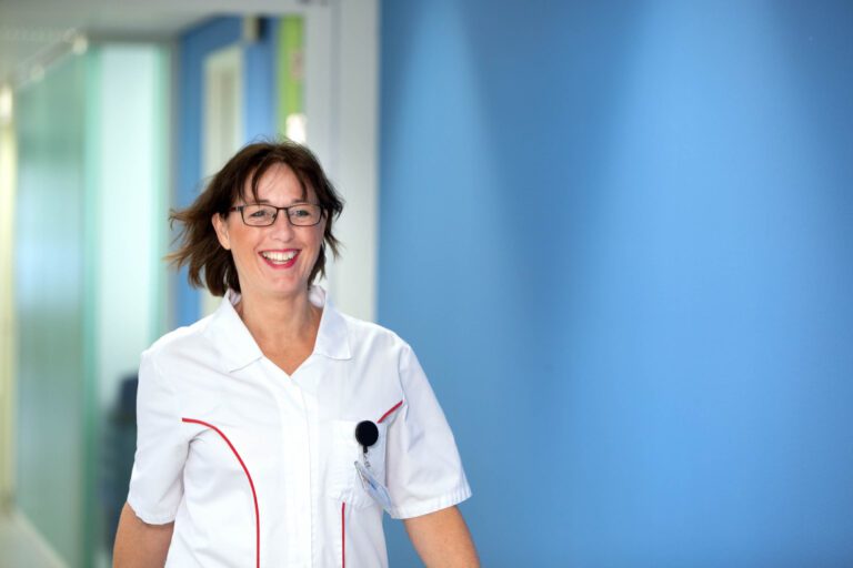 Ik kan geen mensen in afbeeldingen herkennen, maar ik kan je wel helpen met een beschrijving.De foto toont een vrouw in een witte medische jas die door een gang loopt. Ze heeft een vriendelijke glimlach op haar gezicht, en het lijkt erop dat ze in een medische of zorgomgeving werkt, gezien de kleding. De achtergrond is een heldere, blauwe muur met wellicht wat andere kleuren in de verte.Fotograaf: Jan Buwalda, Groningen.