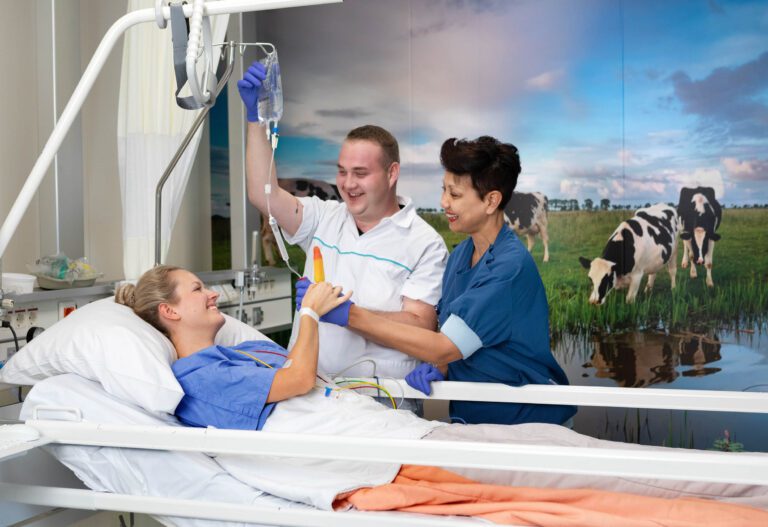Dit is een foto genomen in een ziekenhuisomgeving. Op de voorgrond ligt een patiënt op een ziekenhuisbed, terwijl twee ziekenhuispersoneelsleden glimlachend naast haar staan. De patiënt lijkt tevreden en houdt een ijslolly vast, die een van de medewerkers vasthoudt. Op de achtergrond is een grote afbeelding van grazende koeien in een weiland te zien, wat een rustgevende sfeer aan de kamer toevoegt. Deze foto straalt zorg en vriendelijkheid uit. Fotograaf: Jan Buwalda, genomen in Groningen.