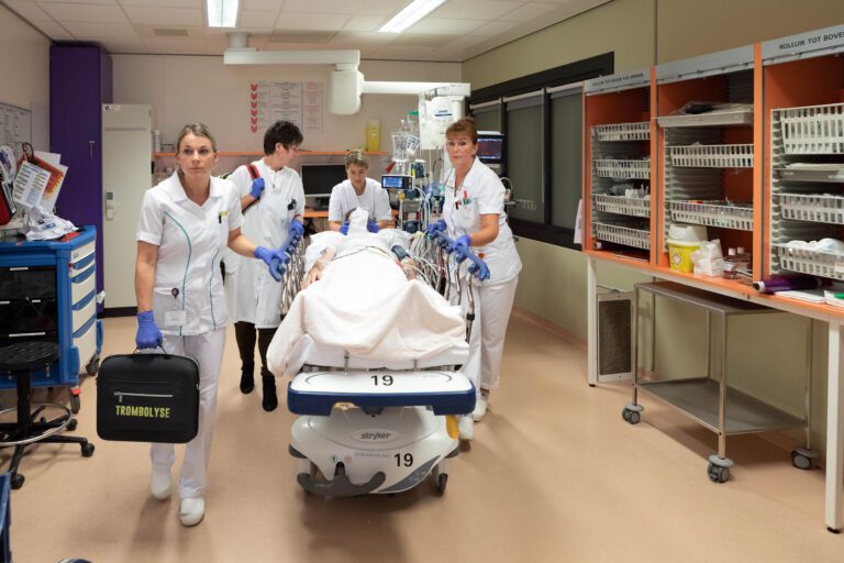 This is a photo showing a busy hospital scene. In the image, four medical professionals in white uniforms are focused and working together, pushing a patient on a hospital bed through a hallway. One of them is carrying a black bag labeled TROMBOLYSE. The setting is well-equipped, with various medical supplies and equipment visible on both sides of the room. The mood conveys urgency and teamwork in a clinical environment.Fotograaf: Jan Buwalda, Groningen.
