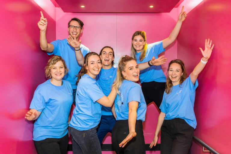 Een groep van zeven mensen, allemaal gekleed in blauwe T-shirts, poseert vrolijk in een felroze ruimte. Ze lachen, hebben hun armen omhoog en lijken zich te vermaken. De sfeer is luchtig en energiek. Foto door Jan Buwalda in Groningen.