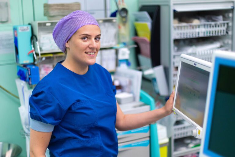 Deze foto, genomen door Jan Buwalda in Groningen, toont een zorgprofessional in een medische omgeving. De vrouw draagt blauwe medische kleding en een paarse muts, en ze staat naast een monitor. De achtergrond laat verschillende medische apparatuur en benodigdheden zien, typerend voor een ziekenhuis of kliniek. Haar houding is ontspannen en vriendelijk, wat een gevoel van vertrouwen en professionaliteit uitstraalt.
