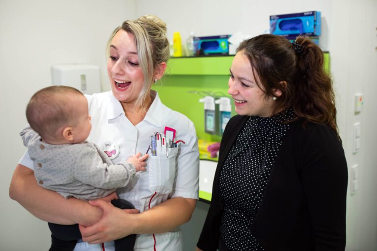 Een vrolijke situatie in een gezondheidszorginstelling in Groningen. Een zorgverlener houdt een baby vast terwijl ze glimlachend met de moeder praat. De zorgverlener draagt een wit uniform met schrijfwaren in de borstzak. Op de achtergrond zijn medische benodigdheden te zien op een felgroene kast. Er heerst een warme en uitnodigende sfeer. Foto door Jan Buwalda.