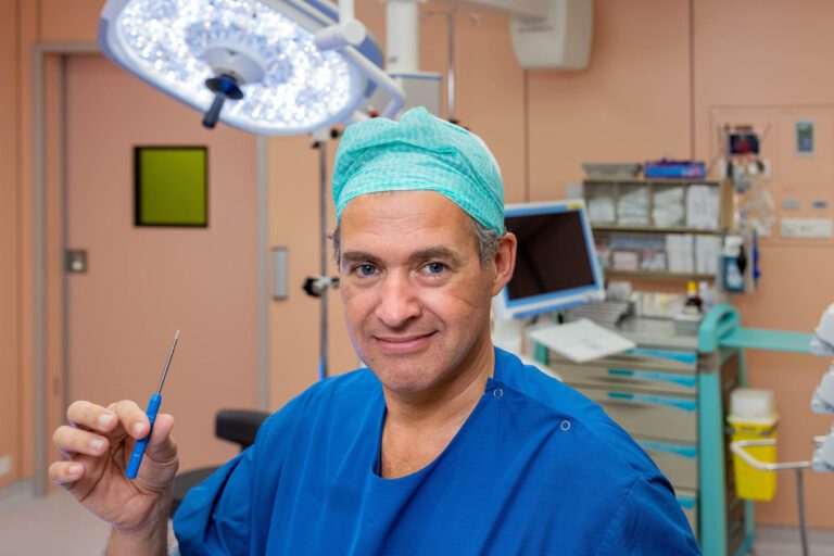 Het beeld toont een chirurg in een operatiekamer, gekleed in een blauwe medische jas en een groene chirurgische muts. De persoon houdt een chirurgisch instrument vast en lijkt klaar voor een procedure. Op de achtergrond zijn medische apparatuur en een operatielamp te zien. De kleurtoon van de kamer is overwegend zacht zalmroze. Fotograaf: Jan Buwalda, Groningen.