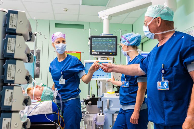 Het is een levendige foto in een operatiekamer, waarop drie medische professionals te zien zijn in blauwe scrubs en chirurgische maskers. Ze lijken bezig met voorbereidingen voor een ingreep. Op de achtergrond is medische apparatuur zichtbaar met schermen die vitale functies monitoren. Een patiënt ligt onder narcose op de operatietafel. De focus ligt op de samenwerking en de precisie van het team. Foto door Jan Buwalda in Groningen.