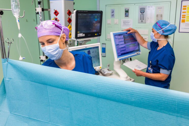 Dit beeld toont twee vrouwelijke gezondheidswerkers in operatiekleding die aan het werk zijn in een medische omgeving. Ze dragen beschermende maskers en haarbedekking. Een van hen concentreert zich op een blauwe chirurgische doek, terwijl de ander iets op een computerscherm controleert. De achtergrond bevat diverse medische apparatuur en monitoren die vitale functies weergeven. De foto is gemaakt door Jan Buwalda in Groningen.