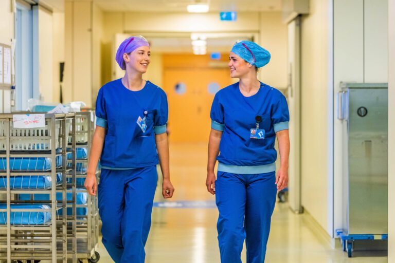 Twee verpleegkundigen lopen samen door een heldere ziekenhuisgang. Ze dragen blauwe uniformen en kleurrijke haarnetjes, terwijl ze kalm en zelfverzekerd met elkaar praten. Aan de zijkant van de gang zijn karren met medische benodigdheden te zien. De sfeer is professioneel en vriendelijk. Foto door Jan Buwalda, Groningen.