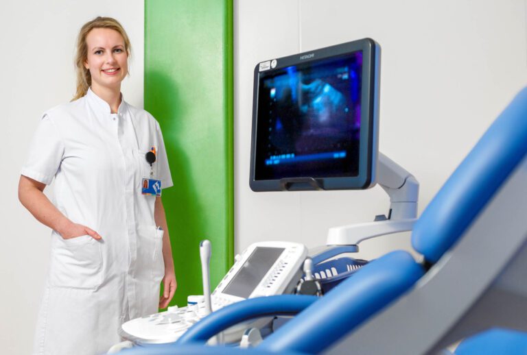 Deze foto, gemaakt door fotograaf Jan Buwalda uit Groningen, toont een medisch professional in een witte uniformjasje. Ze staat met een vriendelijke glimlach naast een echografie-apparaat in een klinische omgeving met een groene muur. Het geheel straalt een gevoel van deskundigheid en vertrouwen uit.