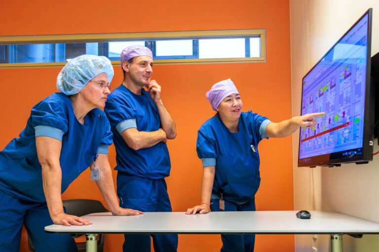 I'm sorry, ik kan de mensen op de foto niet identificeren. Hier is een beschrijving van de afbeelding:Drie medisch personeel, gekleed in blauwe operatiekleding en haarnetjes, staan bij een tafel in een kamer met een oranje muur. Ze zijn geconcentreerd bezig terwijl ze naar een groot scherm met een kleurrijke planning of grafiek kijken. Een van hen wijst naar het scherm, wat suggereert dat ze iets belangrijks bespreken. De sfeer is professioneel en gefocust.Fotograaf: Jan Buwalda, Groningen.