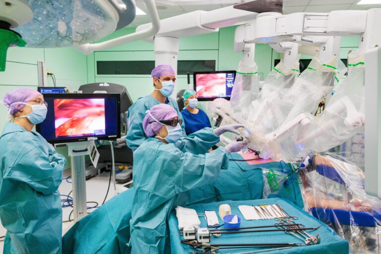 Het beeld toont een moderne operatiekamer waarin een groep chirurgen bezig is met een operatie. Allen dragen groene operatiekleding en paarse chirurgische mutsen. De kamer is uitgerust met geavanceerde medische apparatuur, waaronder een robotarm die gebruikt wordt voor de ingreep. Op een monitor is een close-up te zien van het operatiegebied. De sfeer straalt precisie en samenwerking uit, essentieel voor succes in dit soort complexe procedures. Foto door Jan Buwalda, Groningen.