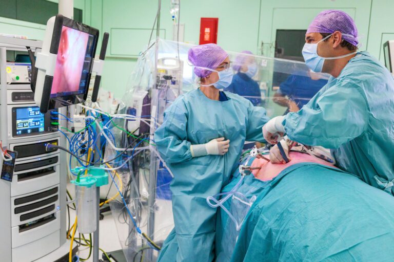 Dit beeld toont een operatiekamer waar een chirurgisch team een ingreep uitvoert. Een arts en een assistent, gekleed in steriele operatiekleding en maskers, werken geconcentreerd aan de patiënt die op de operatietafel ligt. Medische apparatuur en monitoren zijn zichtbaar, en een scherm toont beelden van een endoscopische camera. Foto door Jan Buwalda in Groningen.